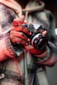 Unrecognizable female photographer wearing stylish coat standing with photo camera in hands on street in evening time against blurred background